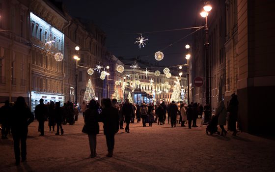 People walking down a snowy city street with festive winter decorations and illuminated buildings at night.