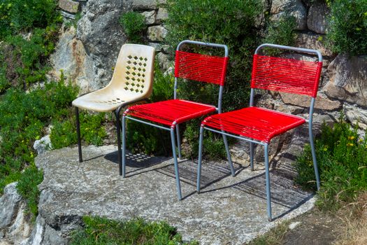 Three colorful chairs on a sunlit rocky terrace surrounded by lush greenery, offering a vibrant outdoor seating arrangement.