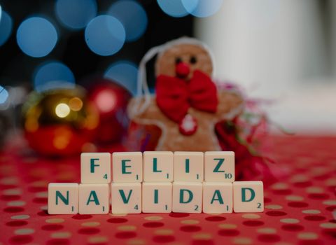 Scrabble tiles spell 'Feliz Navidad' with holiday ornaments and gingerbread man in background.