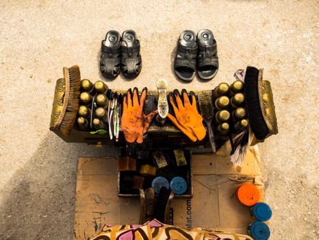 Aerial view of a shoe shine setup with brushes, polish, gloves, and sandals on a concrete surface.