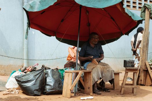 A cobbler repairs shoes under a large umbrella on a sunny day. Urban street scene.