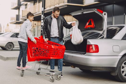 Full body of happy man and boy in casual clothes moving shopping bag from plastic trolley to car trunk on parking near modern buildings