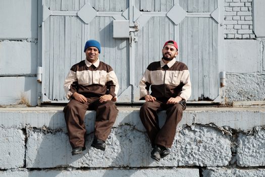 Two industrial workers wearing uniforms sit and smile in front of an old factory door.