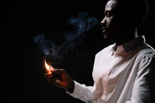 Side view of a man in a white shirt lighting a matchstick with smoke in a dark room.