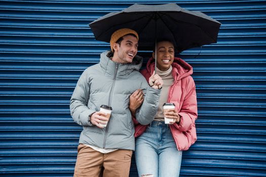Happy multiracial couple with umbrella in warm outerwear laughing while standing on street with takeaway beverages in cold rainy weather