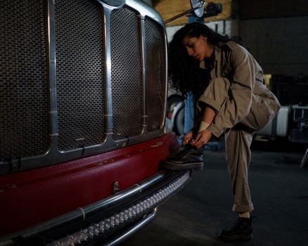 A woman mechanic in coveralls ties her shoes near a truck bumper in a garage setting.