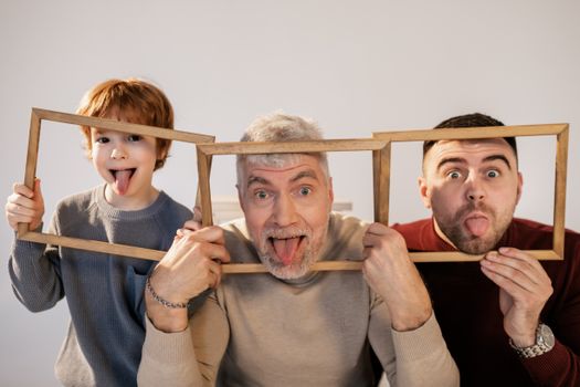 Grandfather, father, and son making funny faces indoors with wooden frames.