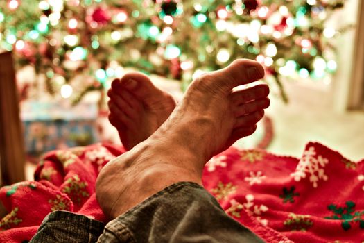 A person relaxing with feet up near a beautifully lit Christmas tree indoors.