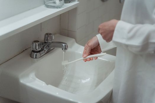 Close-up of a woman washing a toothbrush under a bathroom sink faucet, focusing on dental hygiene